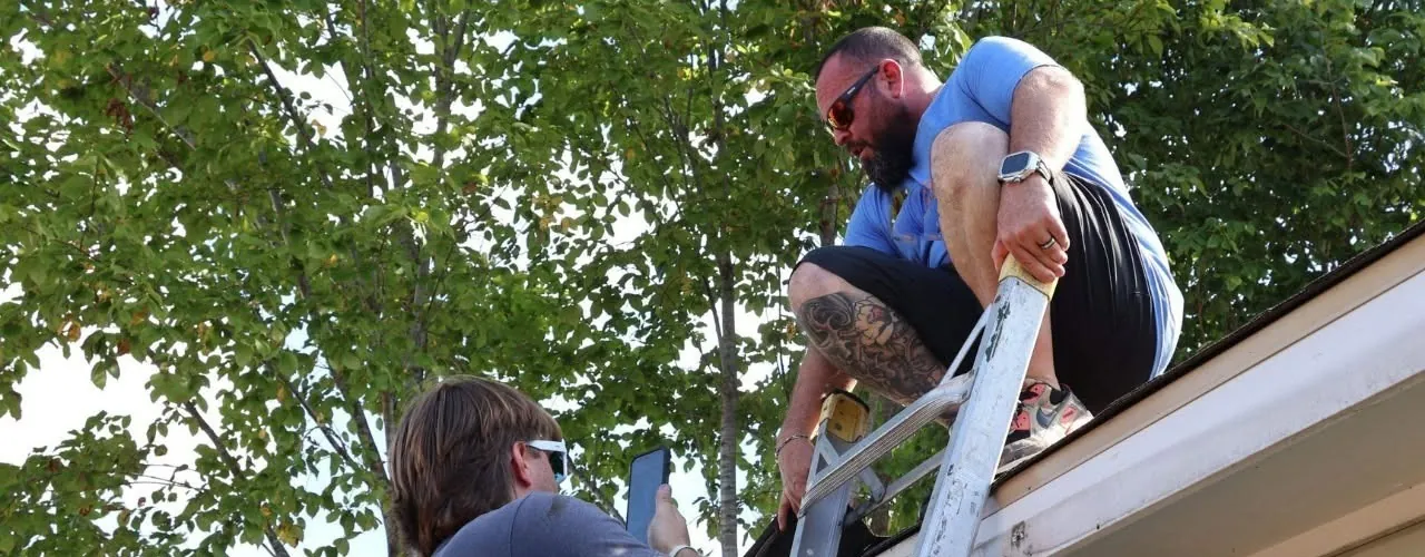 Brian Finley climbing off a ladder onto a roof edge while a crew member holds the ladder with a phone in hand