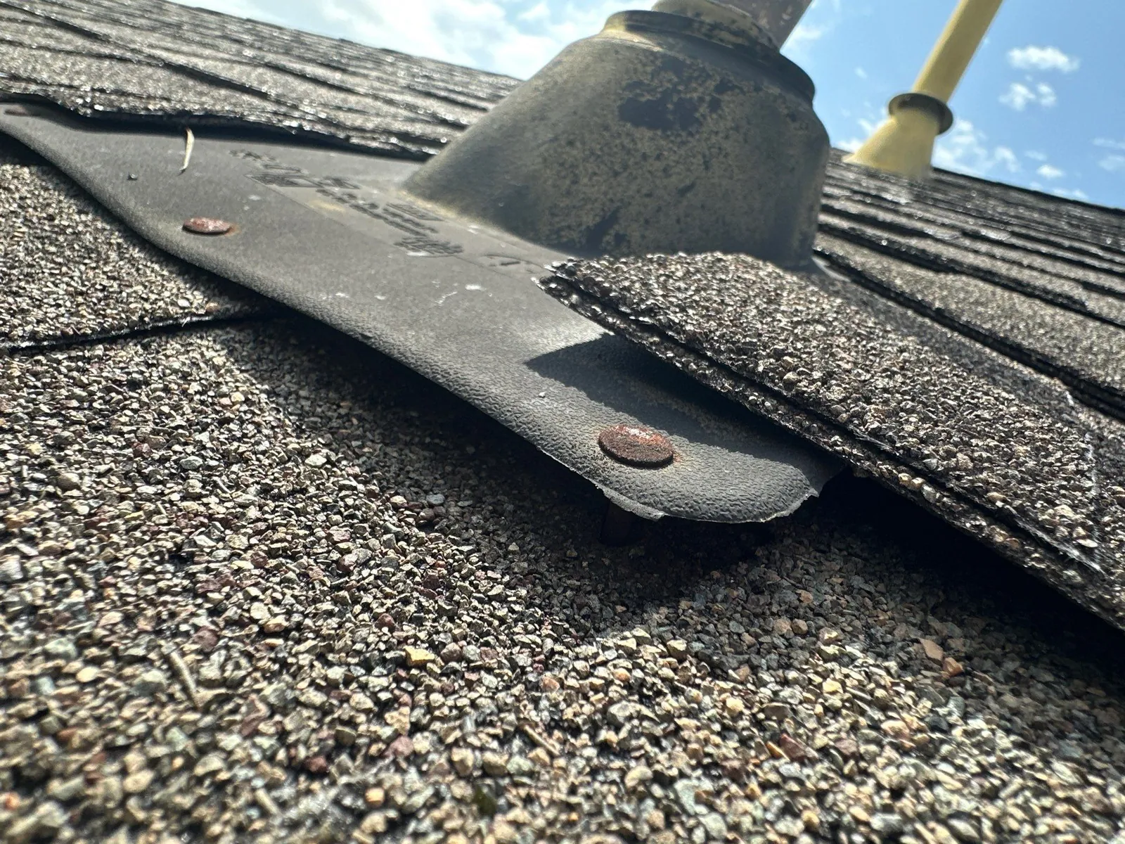 Close-up of a pipe jack flange on a shingle roof, lifting off the shingles where its rusted nail has backed out