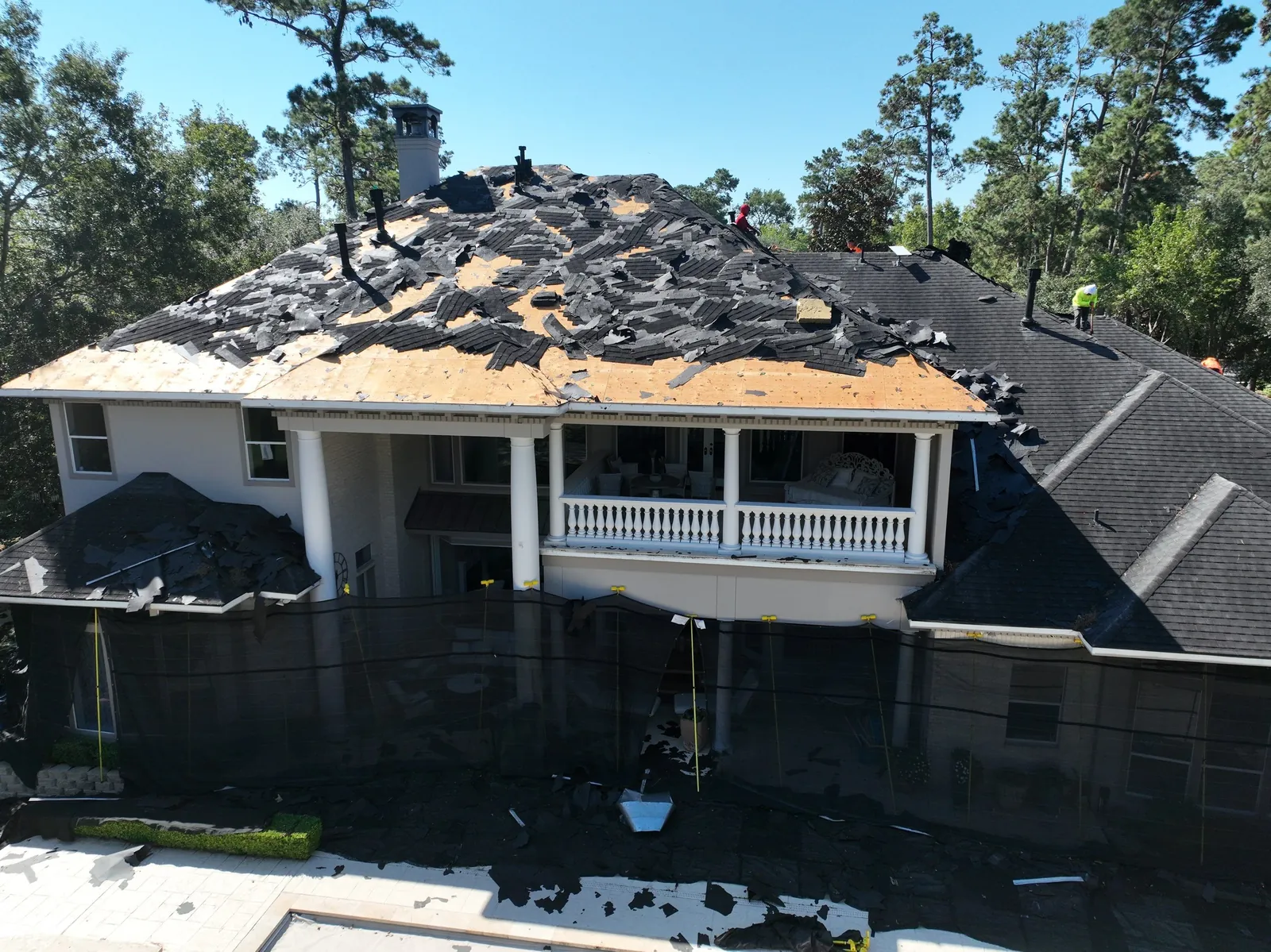 Aerial view of a two-story home mid tear-off, old shingles coming off the deck, black netting hung from the roof edge to the ground
