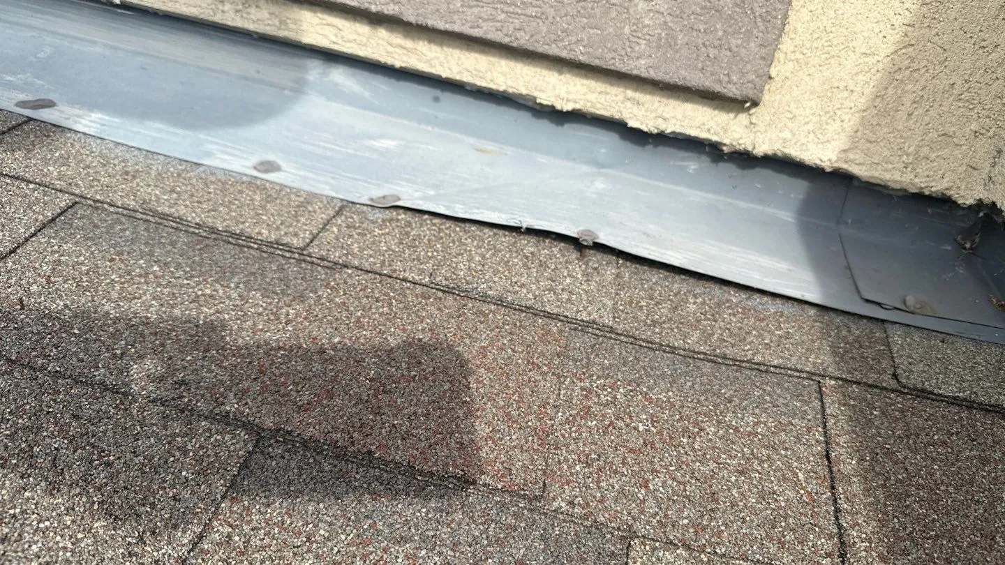 Metal wall flashing where a shingle roof meets a stucco wall, with several nail heads backed out and the flashing edge bent up